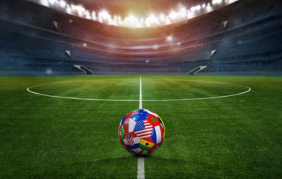 Green textured field in soccer stadium and soccer ball covered with multiple national flags symbolizing an international football tournament.