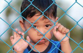 A child refugee stands behind a fence.