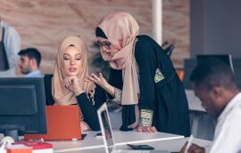 Two women wearing hijabs converse in an office.