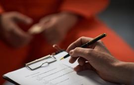 Caucasian woman holding pen and clipboard conducting interview with prisoned adult wearing orange uniform, hands visible in foreground, focus on assessment process.