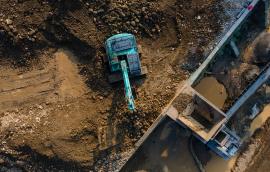  Excavator in Mining Quarry and Rock Crusher Facility on Sunny Day, Industrial mining site captured from above, featuring heavy machinery, material processing operations, rugged excavation terrain