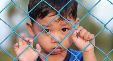 A child refugee stands behind a fence.