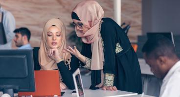 Two women wearing hijabs converse in an office.