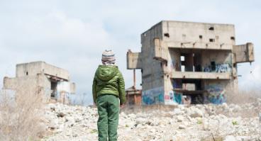 A refugee child looks at ruins of buildings.
