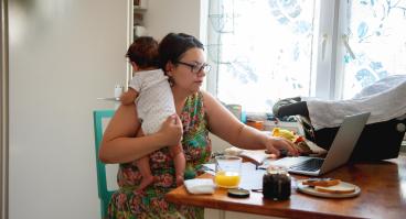 A mother holds her baby while working on her laptop.