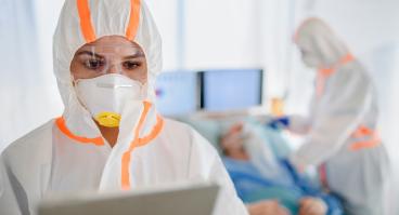 Healthcare workers wear hooded coveralls and masks while interacting with patients.