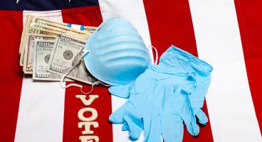 A mask, gloves, money, and letters spelling "vote" lie on a flag of the United States.