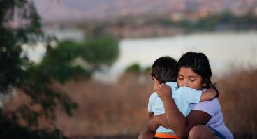 Brother and sister separated from parents are hugging each other in a foreign country, across a river. 