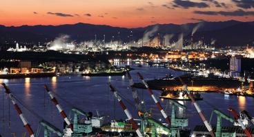 Night view of shipyard with tower cranes near the sea against chimney of oil refinery factory.