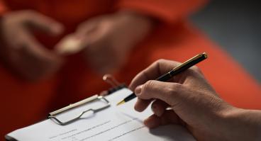 Caucasian woman holding pen and clipboard conducting interview with prisoned adult wearing orange uniform, hands visible in foreground, focus on assessment process.