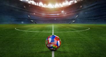 Green textured field in soccer stadium and soccer ball covered with multiple national flags symbolizing an international football tournament.