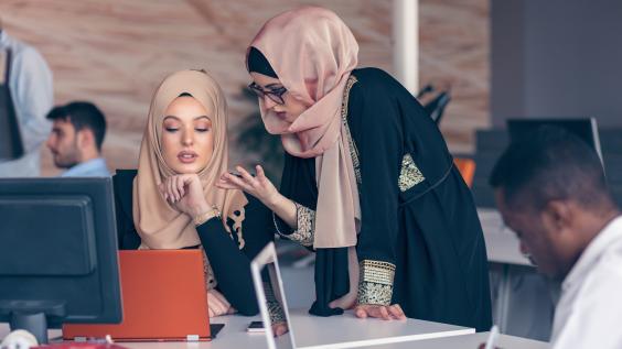 Two women wearing hijabs converse in an office.