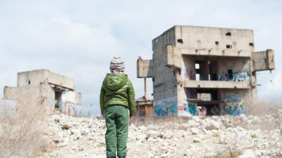 A refugee child looks at ruins of buildings.