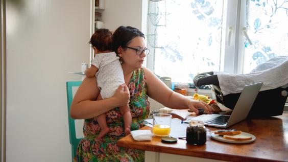 A mother holds her baby while working on her laptop.