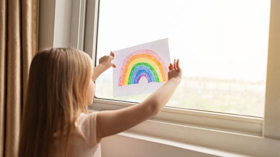 A young child presses a drawing of a rainbow up against a window during quarantine.