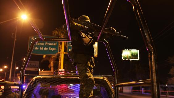 A Mexican police officer bears a rifle while standing on top of a truck.