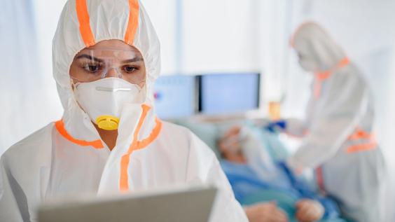 Healthcare workers wear hooded coveralls and masks while interacting with patients.