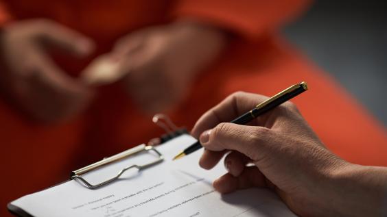 Caucasian woman holding pen and clipboard conducting interview with prisoned adult wearing orange uniform, hands visible in foreground, focus on assessment process.
