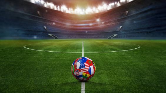 Green textured field in soccer stadium and soccer ball covered with multiple national flags symbolizing an international football tournament.