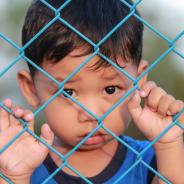 A child refugee stands behind a fence.