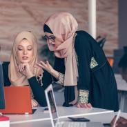Two women wearing hijabs converse in an office.