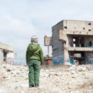 A refugee child looks at ruins of buildings.