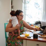 A mother holds her baby while working on her laptop.