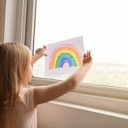 A young child presses a drawing of a rainbow up against a window during quarantine.