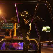 A Mexican police officer bears a rifle while standing on top of a truck.