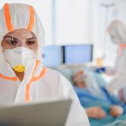 Healthcare workers wear hooded coveralls and masks while interacting with patients.