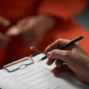 Caucasian woman holding pen and clipboard conducting interview with prisoned adult wearing orange uniform, hands visible in foreground, focus on assessment process.