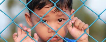A child refugee stands behind a fence.