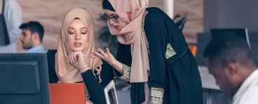 Two women wearing hijabs converse in an office.