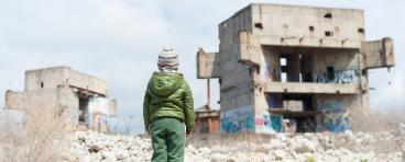 A refugee child looks at ruins of buildings.