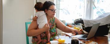 A mother holds her baby while working on her laptop.