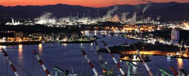Night view of shipyard with tower cranes near the sea against chimney of oil refinery factory.