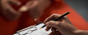 Caucasian woman holding pen and clipboard conducting interview with prisoned adult wearing orange uniform, hands visible in foreground, focus on assessment process.